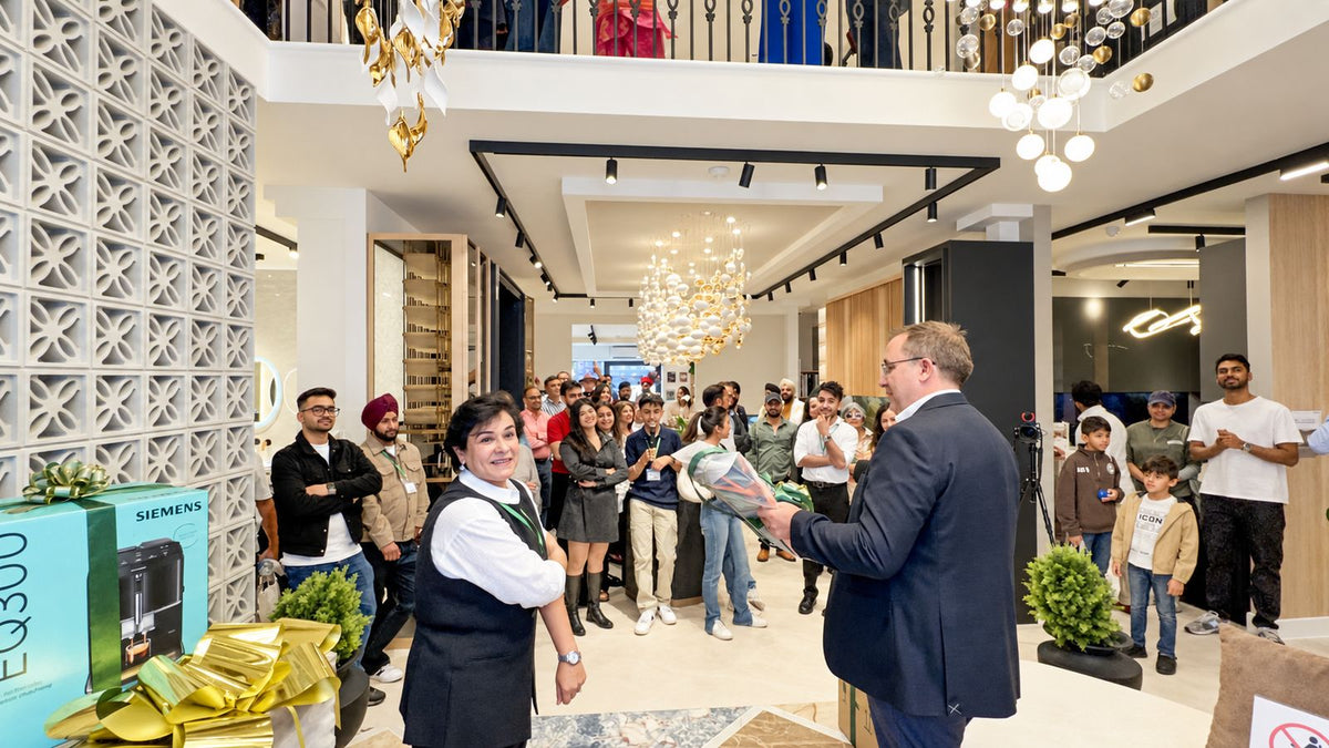 A group of people gathered inside a modern showroom or retail space, with a man in a navy suit presenting a gift box with a gold ribbon to a woman in a dark vest and white shirt. A Siemens product box is visible on the left, and the background features decorative walls, chandeliers, and a staircase.
